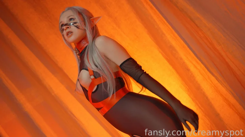 Busty Creamy Spot in elf ears and orange harness, posing in black bodysuit against orange backdrop