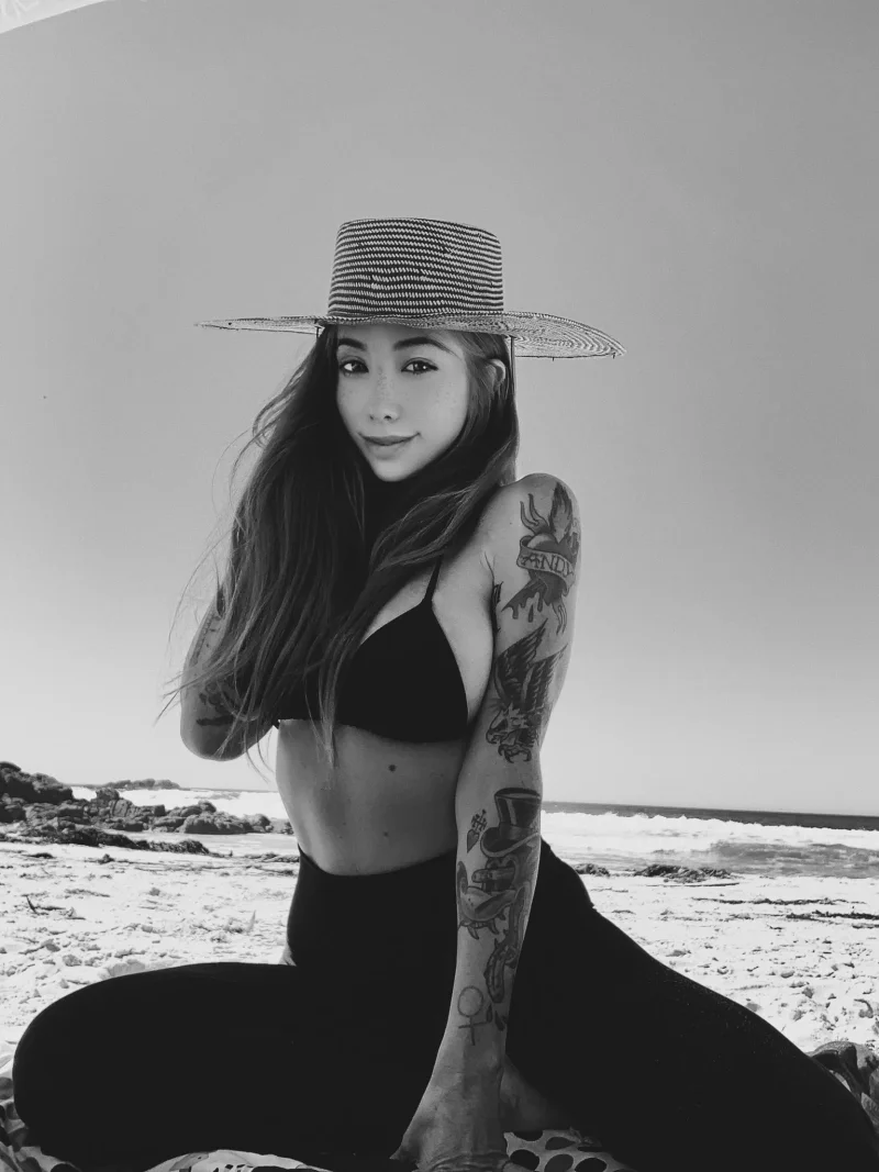 Naughty Coconut Kitty in black bikini top and pants, sitting on beach with tattoos showing